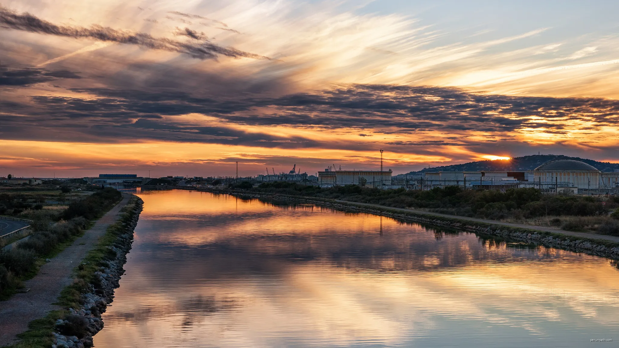The lights of the end of the day reflected in a canal in Frontignan. In the background the Refinery of Frontignan and the Mount Saint-Clair in Sète. | Autor: Christian Ferrer | Licencja: CC BY 4.0 | Źródło: Wikimedia Commons