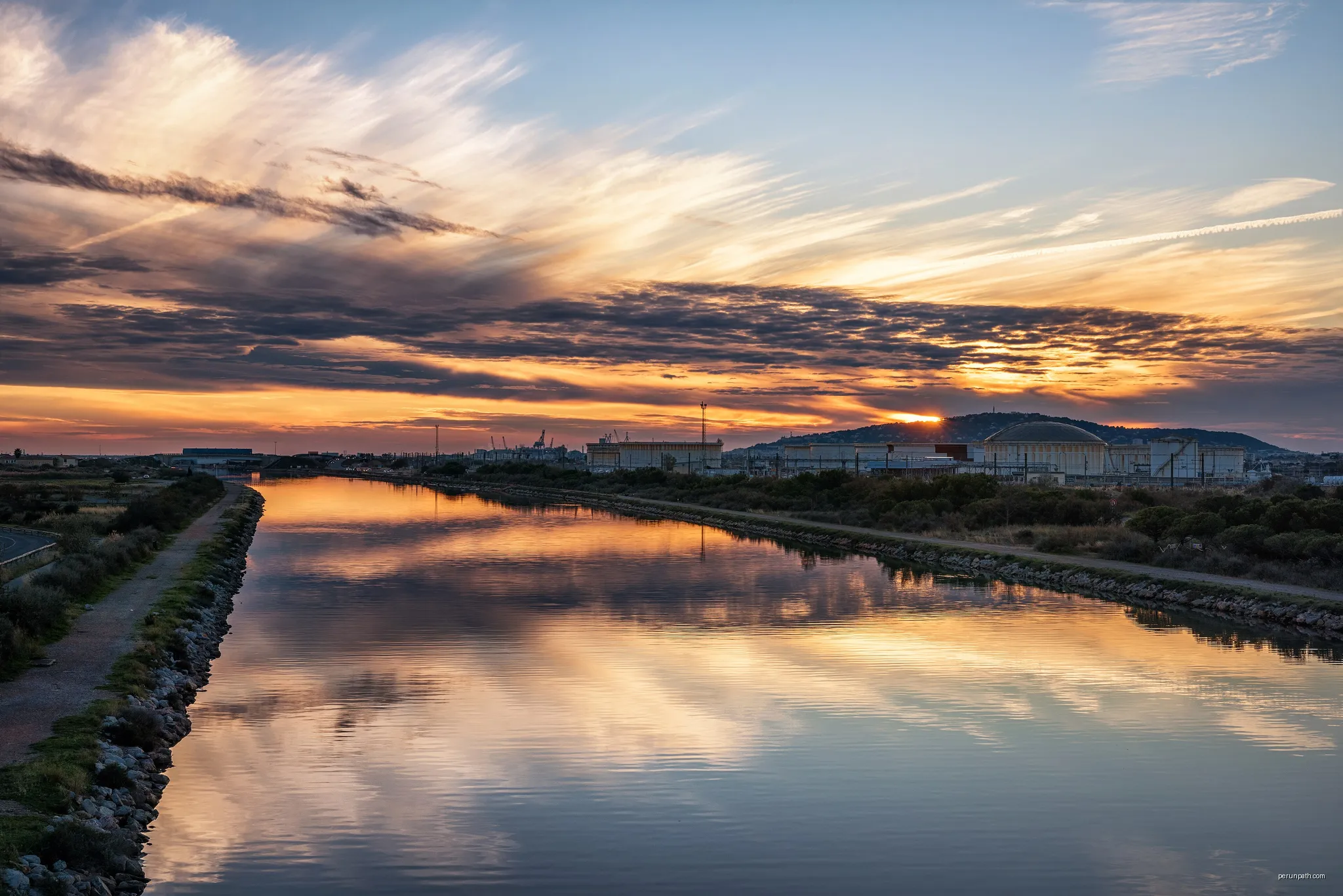 The lights of the end of the day and of the sunset reflected in a canal in Frontignan. On the right in the background the Refinery of Frontignan and the Mount Saint-Clair in Sète. | Autor: Christian Ferrer | Licencja: CC BY 4.0 | Źródło: Wikimedia Commons