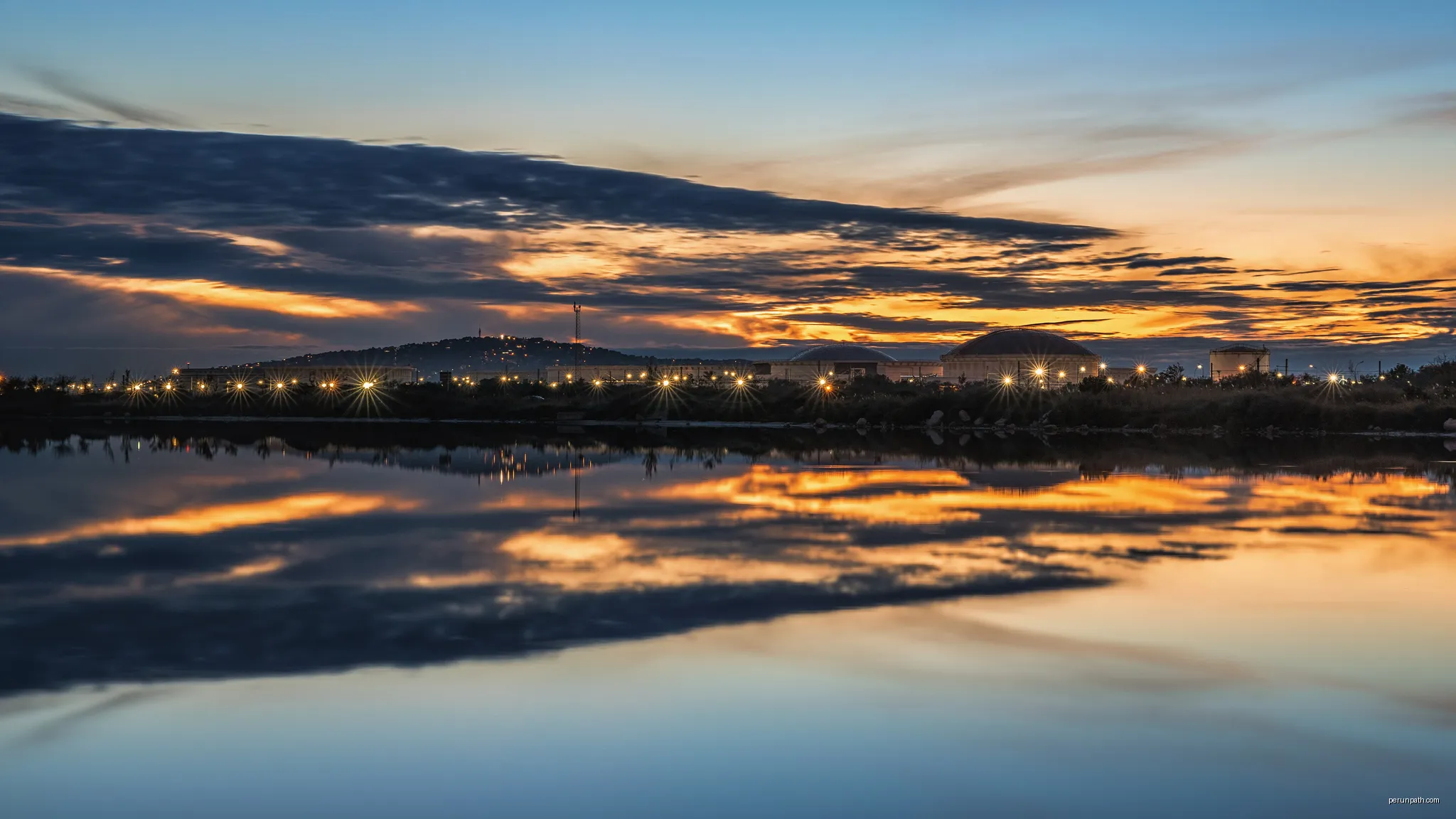 The lights of the end of the day reflected in a canal in Frontignan. In the background on the left the Mount Saint-Clair in Sète, and on the right the Refinery of Frontignan. | Autor: Christian Ferrer | Licencja: CC BY 4.0 | Źródło: Wikimedia Commons